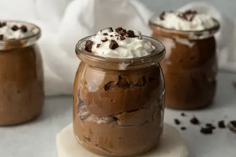 Czekoladowy pudding proteinowy w słoiczku, udekorowany bitą śmietaną i kawałkami czekolady.