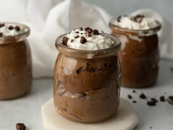 Czekoladowy pudding proteinowy w słoiczku, udekorowany bitą śmietaną i kawałkami czekolady.